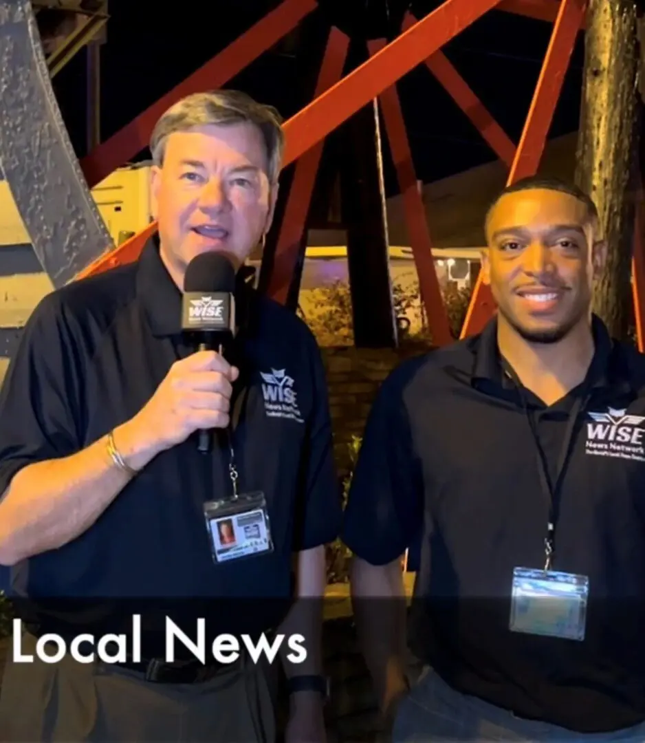 Two news reporters stand outdoors at night, one holding a microphone, both wearing black shirts with badges. A structure with red beams is in the background. The caption reads "Local News.