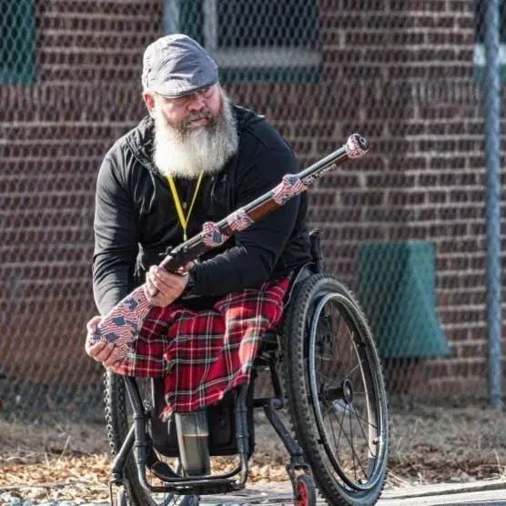 A man with a gray beard in a wheelchair holds a decorated rifle. He wears a cap, black jacket, red plaid kilt, and has a yellow lanyard around his neck. He is in front of a brick building with a chain-link fence.