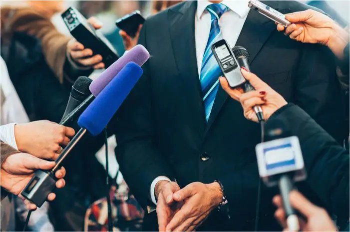 A person in a suit is surrounded by journalists holding microphones and recording devices, suggesting a press conference or media interview. The face is not visible.