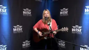 A woman with long blonde hair is playing an acoustic guitar and singing into a microphone in a studio with WISE News Network banners in the background.