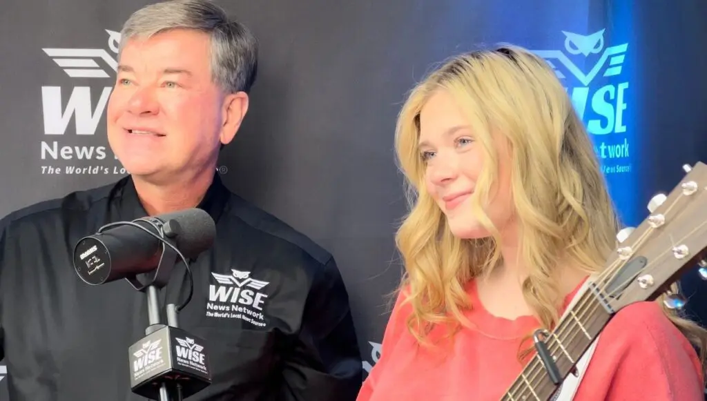 A man and a woman stand together in front of microphones. The man is wearing a black shirt with the logo "WISE News Network," and the woman, holding a guitar, is wearing a red top.