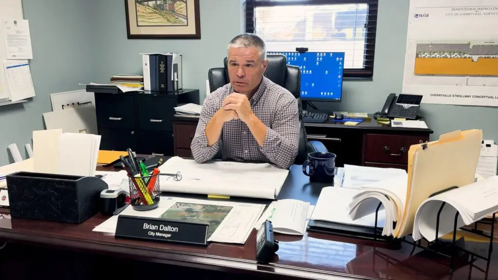 A man sits at a desk in an office with paperwork, a computer, and a nameplate that reads "Brian Dalton, City Manager.