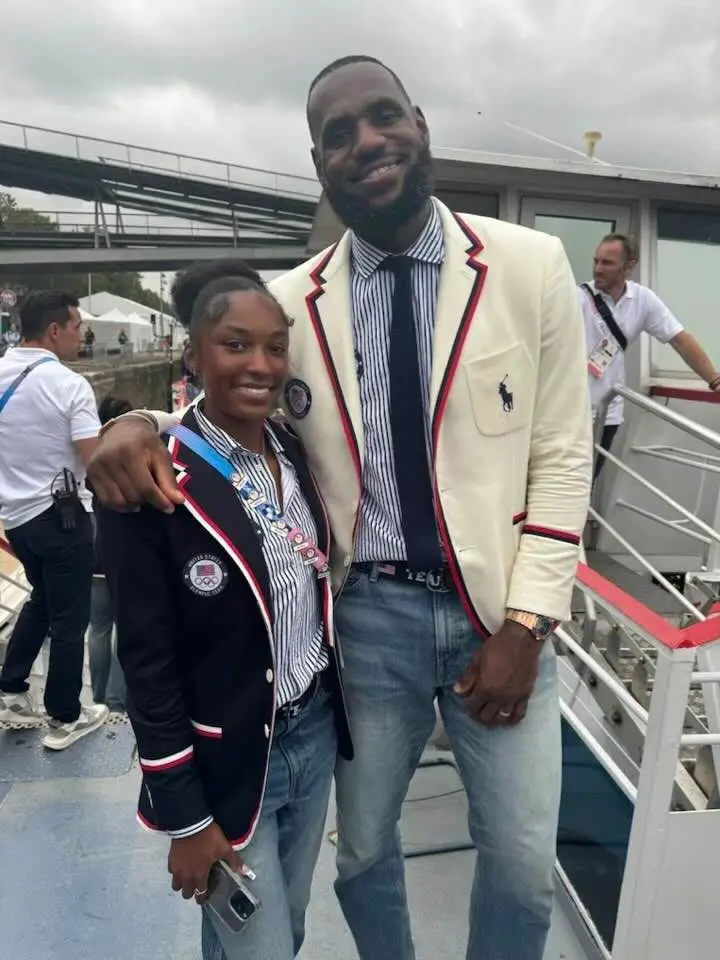 Two people stand beside each other, both wearing matching blazers and jeans. The taller person is smiling with an arm around the shorter person, who is also smiling. A dock and bystanders are in the background.