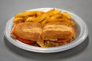 A plate with a sesame seed bun cheeseburger topped with tomatoes, lettuce, and cheese, accompanied by a serving of crinkle-cut fries.