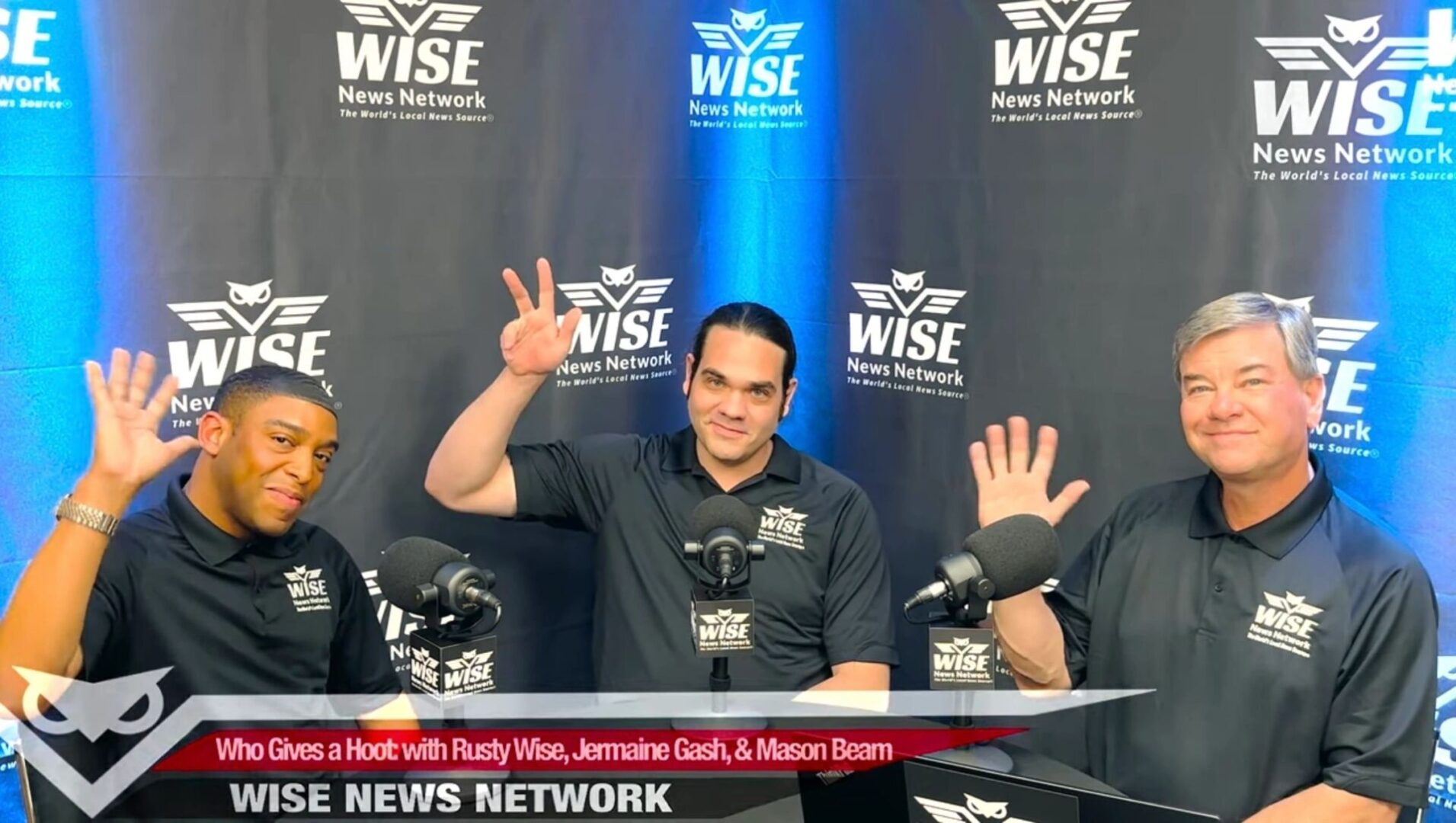 Three individuals seated at a news desk with microphones smile and wave. The backdrop displays "WISE News Network." A banner reads "Who Gives a Hoot with Rusty Wise, Jermaine Gash, & Mason Beam.