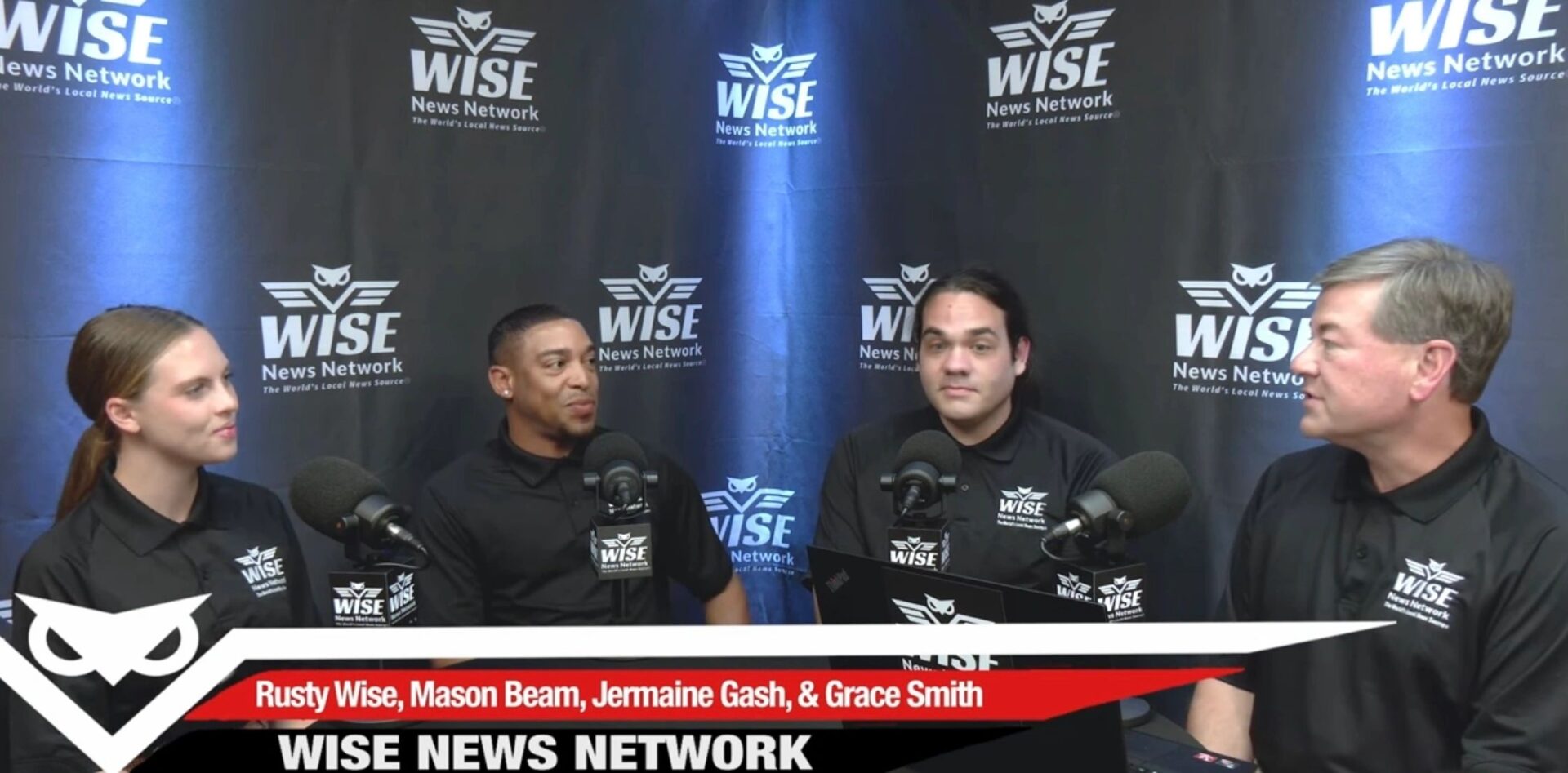 Four individuals in black shirts are seated in front of microphones with "WISE News Network" banners in the background. They are participating in a discussion panel for the WISE News Network.