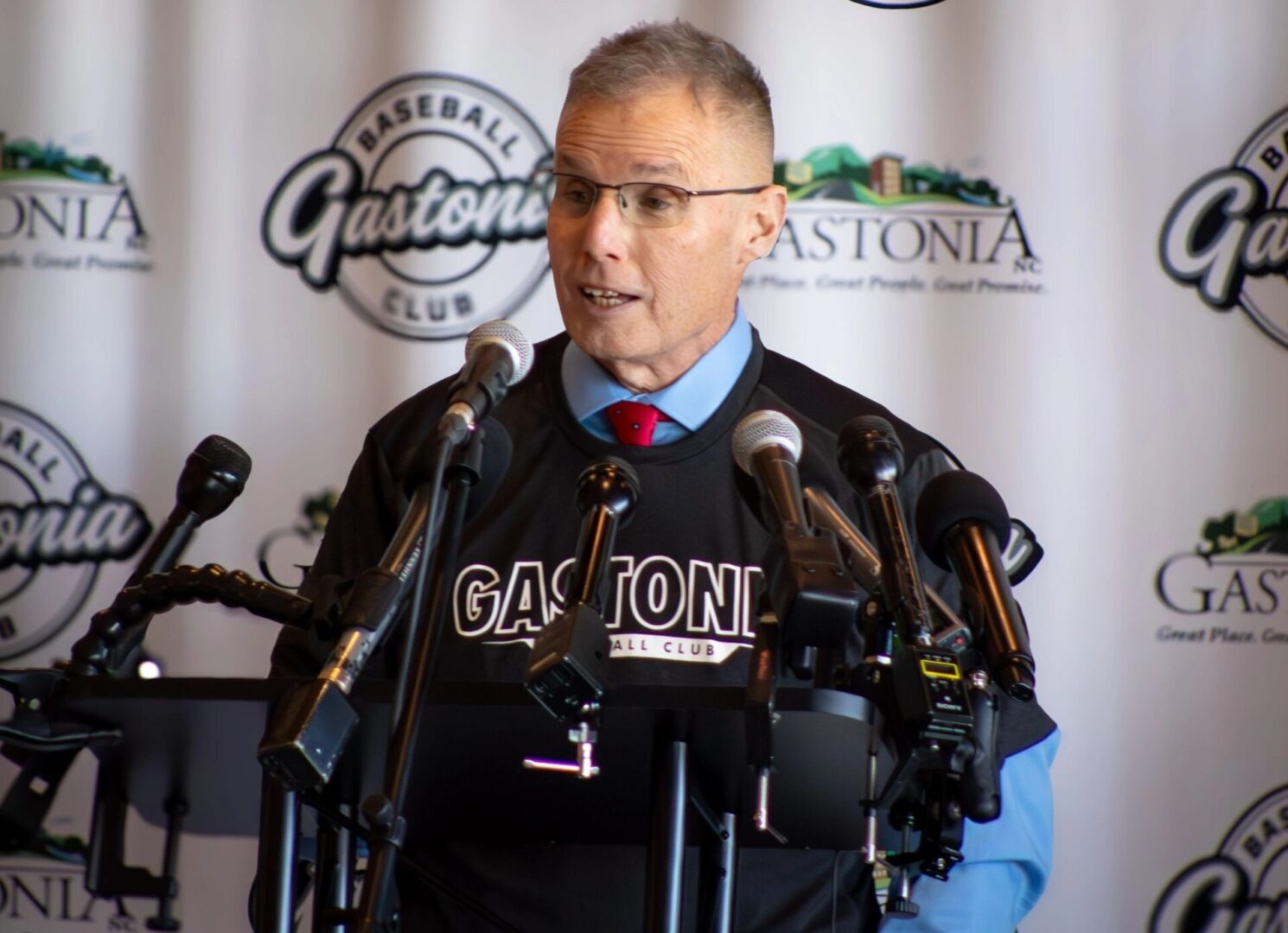 A man in a Gastonia Baseball Club shirt speaks at a podium surrounded by microphones with a Gastonia backdrop behind him.