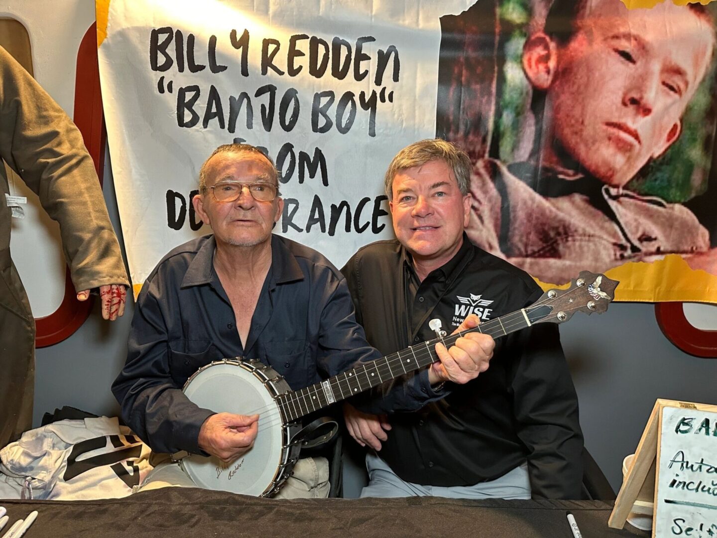 Two men seated at a table pose with a banjo. A banner behind them reads "Billy Redden 'Banjo Boy' from Deliverance" with a portrait of one of the men in a scene from the film.