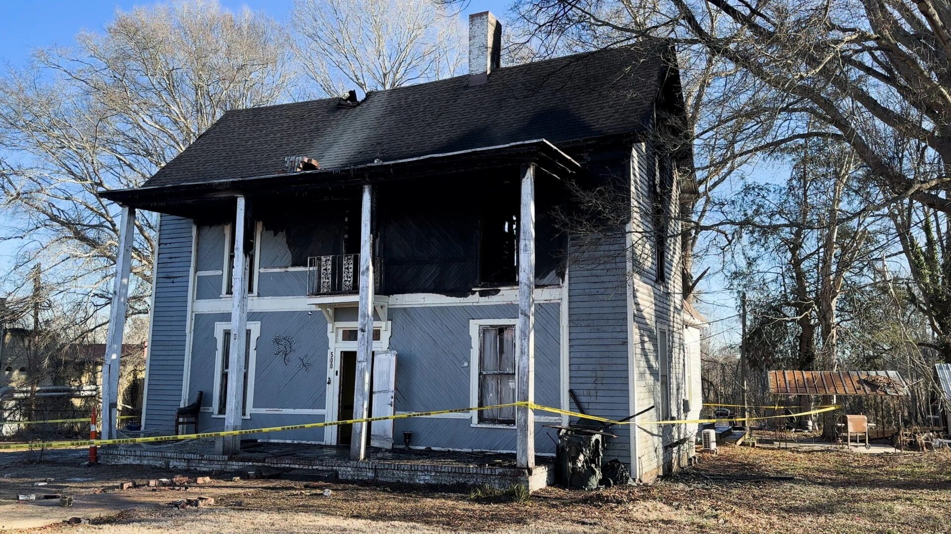 A two-story house with significant fire damage is surrounded by yellow caution tape. The exterior shows signs of charring and structural damage, and the windows are visibly broken or boarded up.