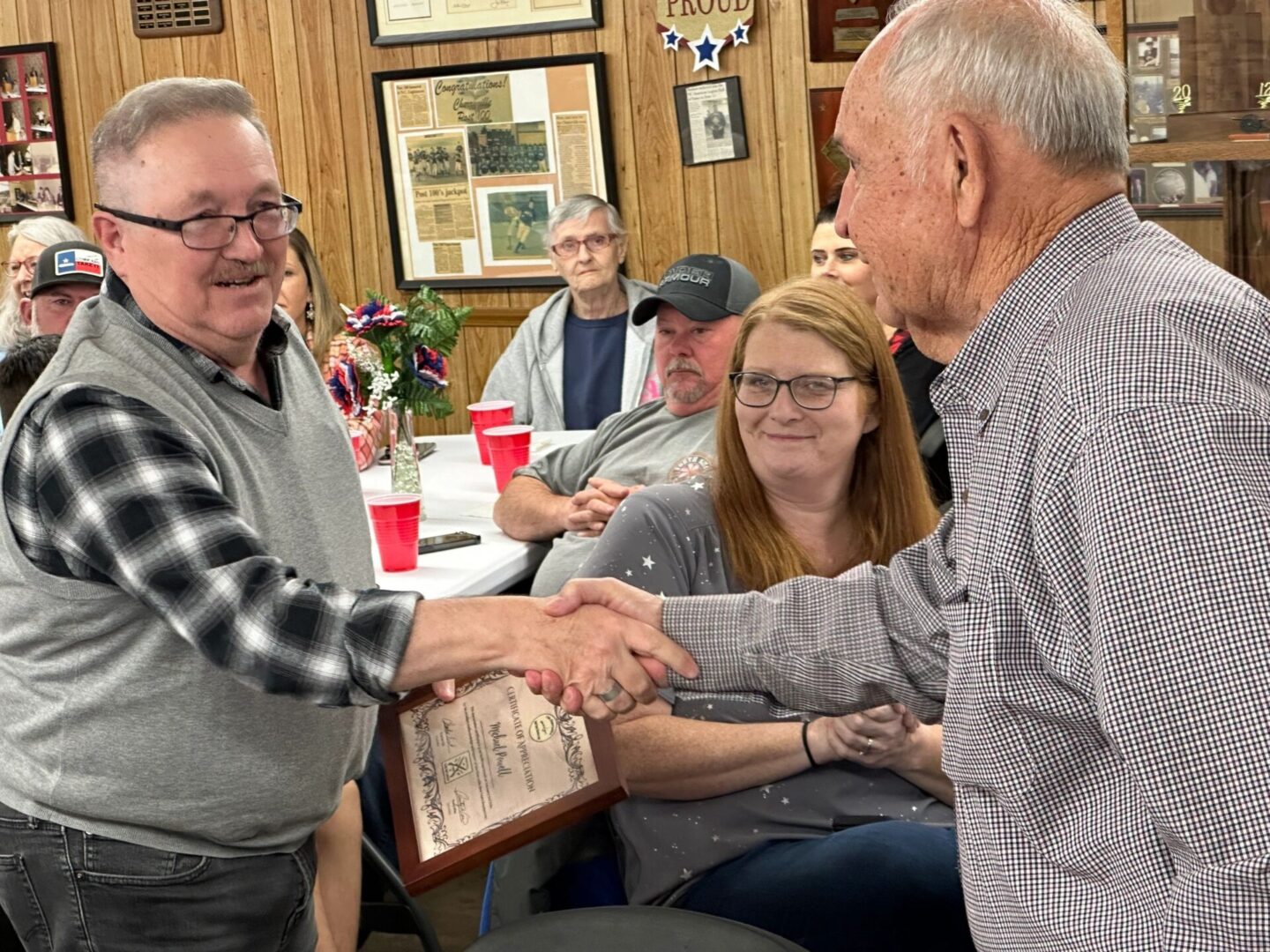 Two men are shaking hands while one of them holds a plaque. Several people are seated at tables in the background, observing the interaction.