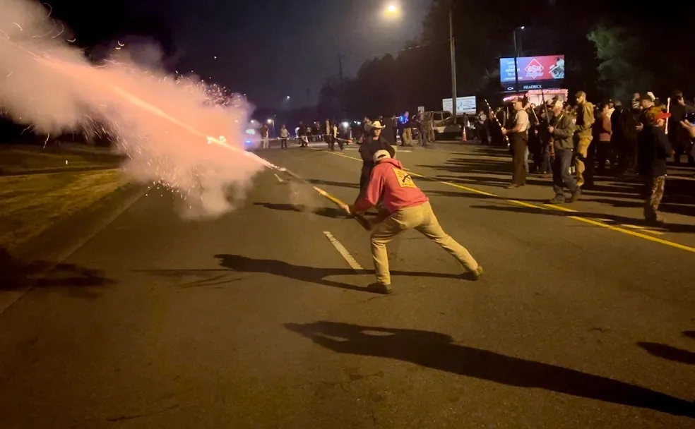 A person launches a firework in the direction of a group of people on a street at night. The crowd stands watching, with smoke illuminated by the streetlights.