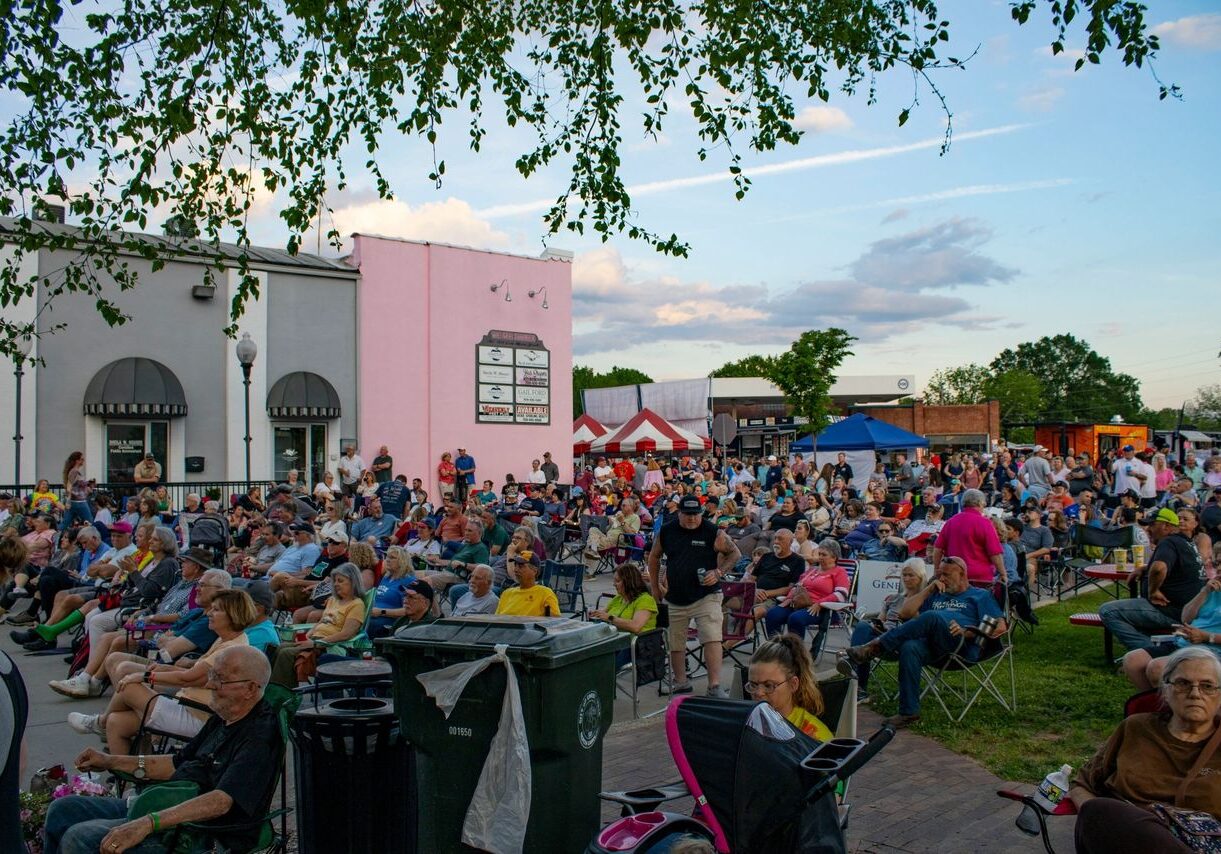 Large outdoor gathering with people seated on chairs and blankets in a town square, surrounded by trees, buildings, and food stalls. The sky is partly cloudy.