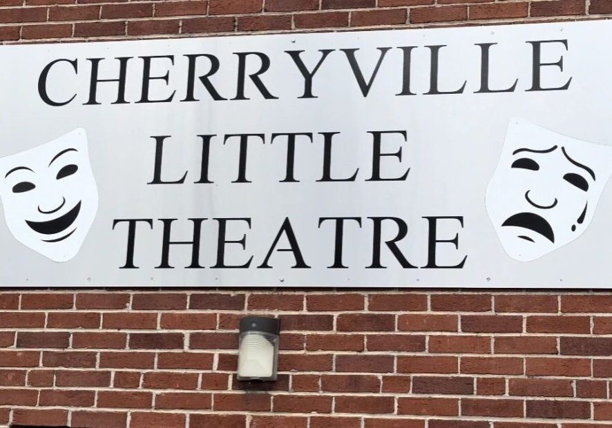 A sign on a brick wall reads "Cherryville Little Theatre" with happy and sad theatrical masks on either side.