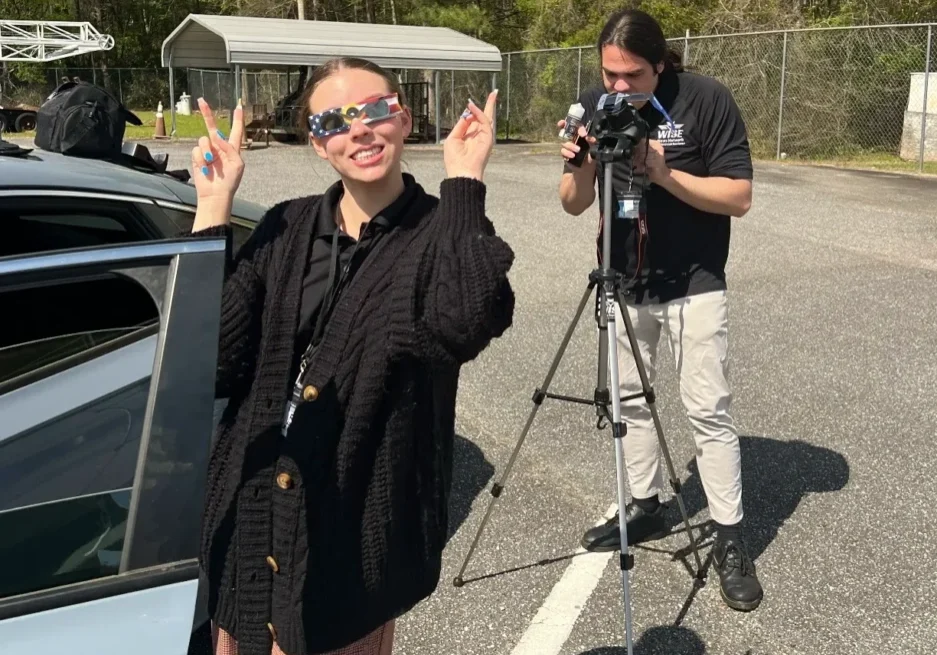 A person stands beside a car making a peace sign with both hands while another person operates a camera on a tripod nearby in an outdoor setting.