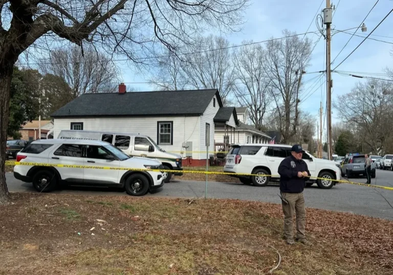 Law enforcement vehicles and an officer are present outside a house with yellow crime scene tape surrounding the area.