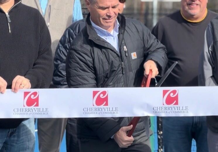 A group of men stand behind a ribbon labeled "Cherryville Chamber of Commerce" at a ribbon-cutting ceremony. One man in the center is cutting the ribbon with a large pair of scissors.