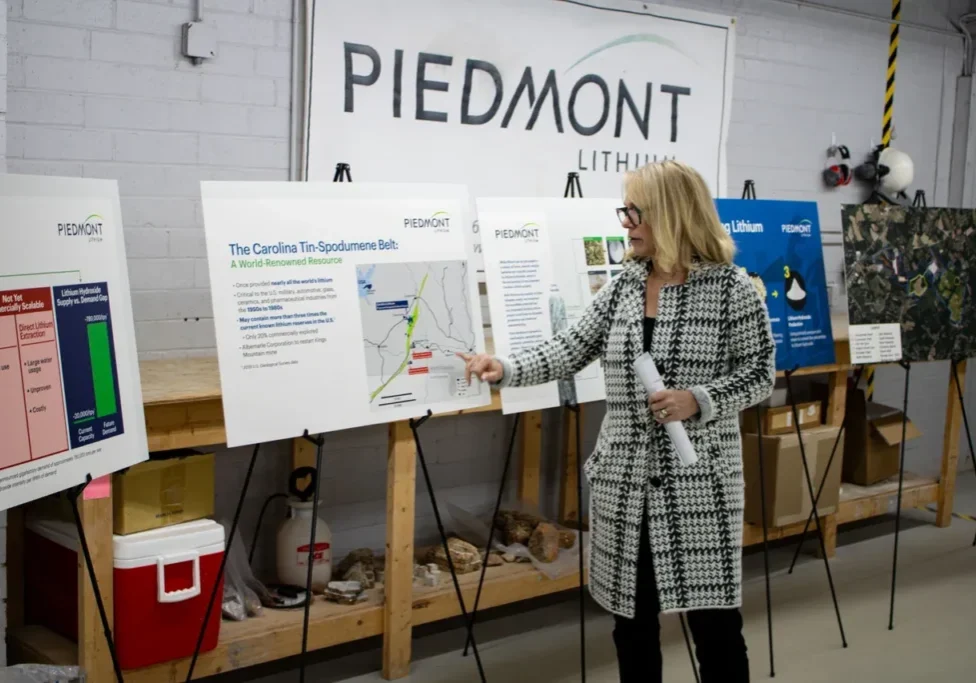 A person stands next to informational posters at a presentation for Piedmont Lithium, pointing towards a map showing the Carolina Tin-Spodumene Belt. They are holding rolled-up papers.