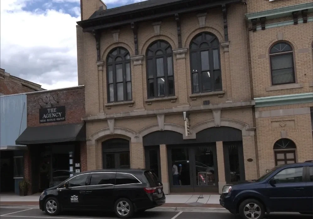 A street view of brick buildings, including a business labeled "The Agency Real Estate Group". Several parked cars and a cloudy sky are also visible.