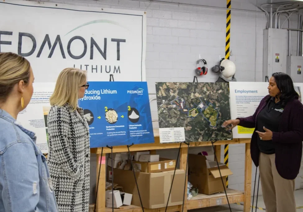 Three people in a room observe a display on Piedmont Lithium's operations. One person gesturing towards the display, with multiple posters on a table and a company banner on the wall.