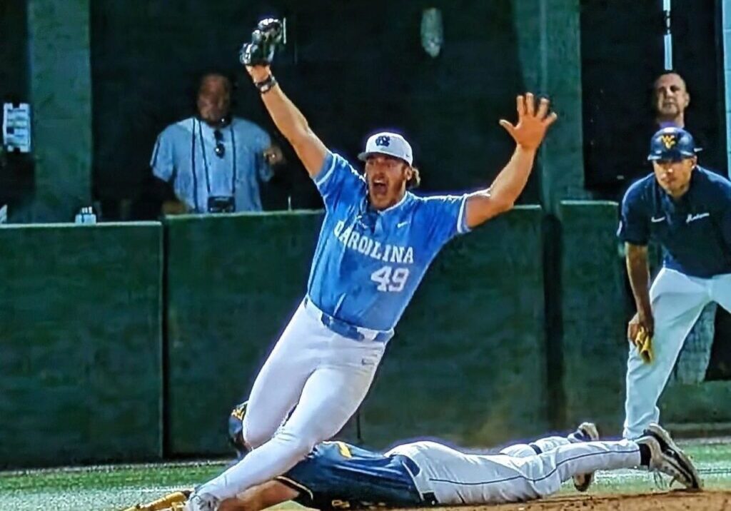 Baseball player in a Carolina jersey (#49) energetically signals an out while another player slides into a base. Two people watch from the background.