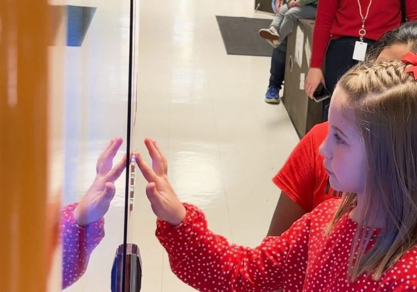 A young girl in a red polka-dot shirt touches a display screen with her hand while standing in a hallway. Another person stands behind her.