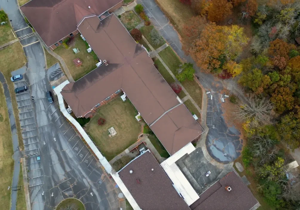 Aerial view of a large building with a brown roof surrounded by parking lots, grassy areas, and autumn trees. Paths and walkways are visible around the property.