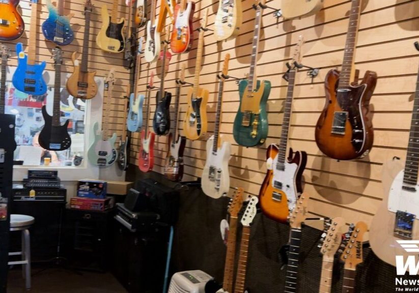 A music store displays a variety of electric guitars and basses on slatwall panels. Amplifiers and other accessories are seen in the lower part of the image. A "WISE News Network" logo is in the bottom right.