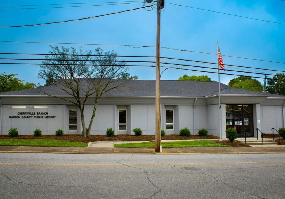 A gray building with a sign that reads "Cherryville Branch Gaston County Public Library," a few shrubs, an American flag, and a tree in front, viewed from across the street.