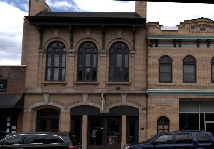 A two-story brick building with arched windows, adjacent to a building with "The Agency Real Estate Group" sign, against a cloudy sky.