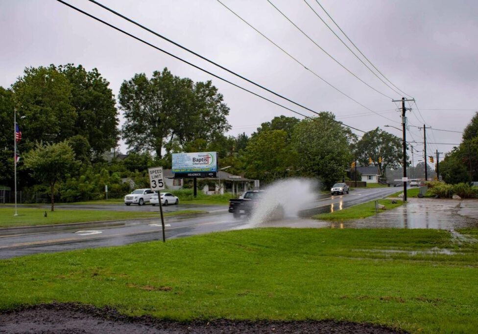 A car drives through a large puddle on a wet road, creating a splash. The sky is overcast, and there are other vehicles and greenery around. A speed limit sign and billboard are visible.