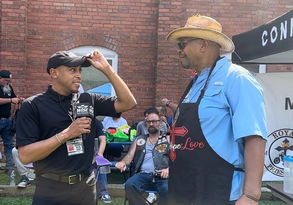 Two men are having a conversation outdoors. One is holding a microphone and wearing a black shirt and cap; the other wears a straw hat, blue shirt, and a black apron with a heart design.