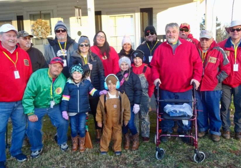 A group of people, including children and adults, pose outdoors in front of a house. Many are dressed warmly, holding rifles, with one person using a walker.