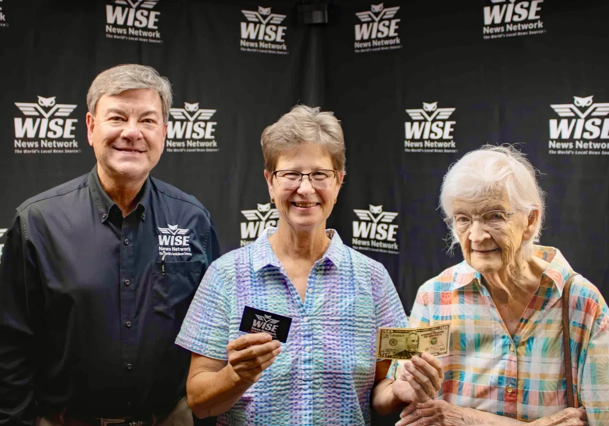 Three people are standing in front of a WISE News Network backdrop. Two are holding a WISE card and one is holding a twenty-dollar bill.