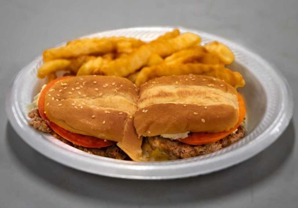 A plate with a sesame seed bun cheeseburger topped with tomatoes, lettuce, and cheese, accompanied by a serving of crinkle-cut fries.