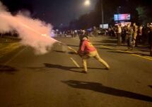 A person in a red shirt and white helmet directs a stream of red smoke during a nighttime street protest with onlookers in the background.