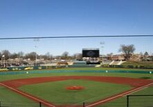 A well-maintained baseball field with a clear sky, featuring an infield with bases, green outfield, and a large scoreboard in the background. Stadium seating surrounds the field.